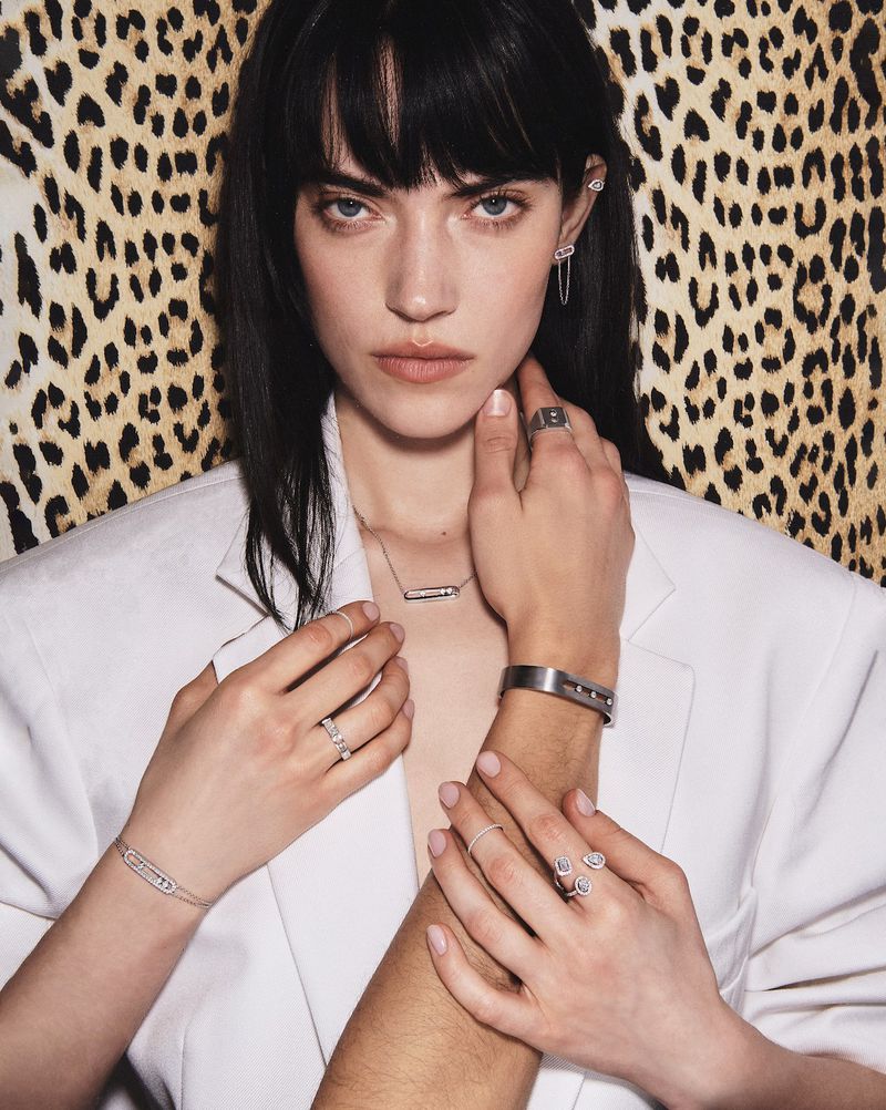 Portrait of a woman with black hair wearing a white jacket and jewelry, posing with a man's arm, against a leopard-print background.