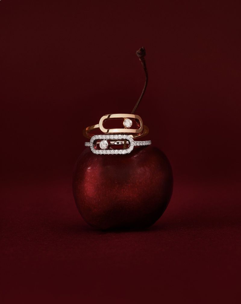 Two gold and diamond rings rest on a deep red cherry, studio lighting against a deep red background, jewelry product shot.