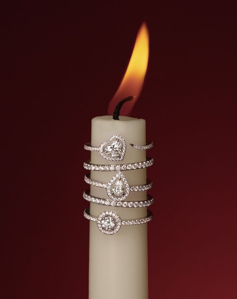 Close-up of sparkling diamond rings of varied shapes stacked on a lit candle against a dark red background, jewelry advertisement.