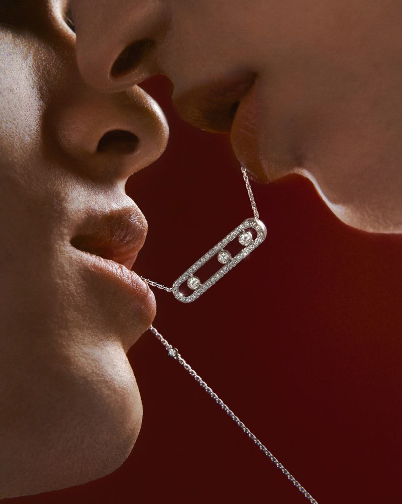 Close-up of two people holding a diamond necklace in their mouths, set against a red backdrop for a luxury jewelry advertisement.