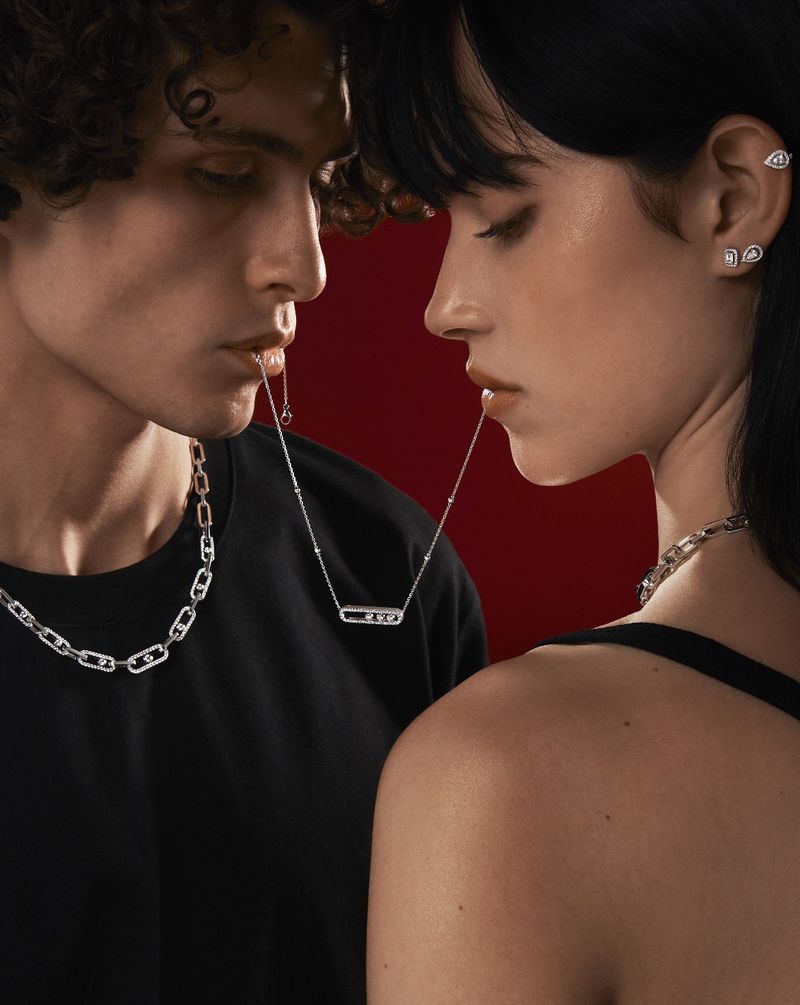 Two young adult fashion models showcase fine jewellery. Man and woman are posing in a studio against a red backdrop with silver necklaces.