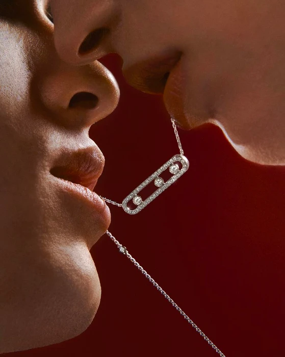 Close-up of a man and a woman holding a diamond pendant necklace between their lips against a red background.
