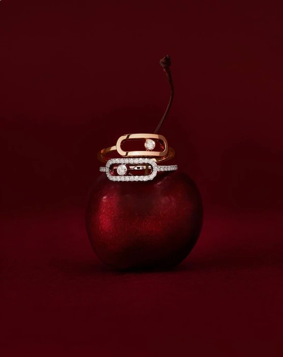 Two diamond and gold rings resting on a glossy red cherry against a dark crimson studio background.