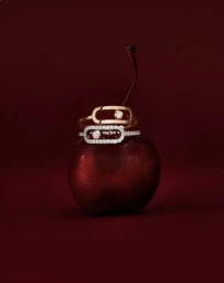Two diamond and gold rings resting on a glossy red cherry against a dark crimson studio background.