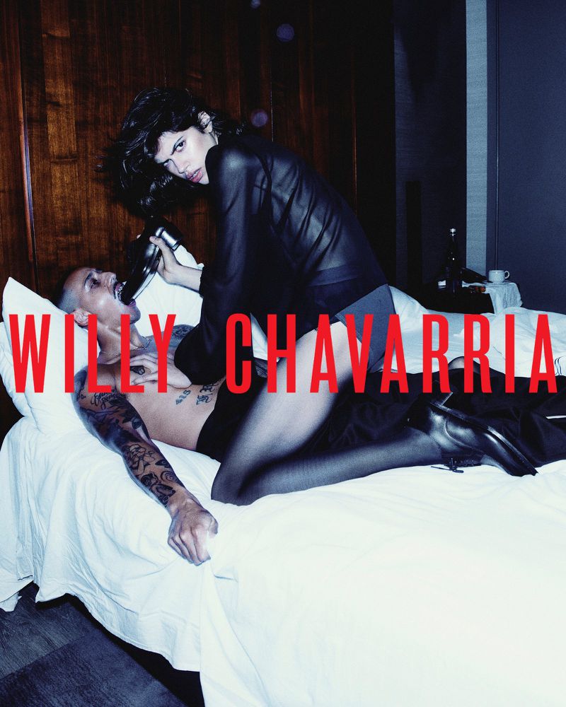 Model kneeling over a man lying on a bed holding a dress shoe in his mouth in a hotel room