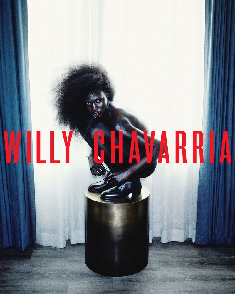 Model crouching on a dark metallic pedestal wearing black shoes in a room with blue curtains and a window backdrop