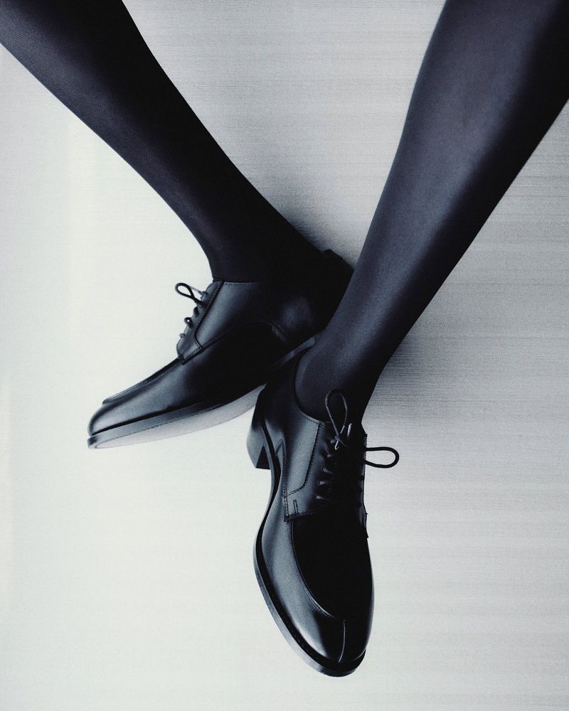 Close-up of shiny black leather dress shoes paired with dark tights, set against a simple grey backdrop in a studio setting.