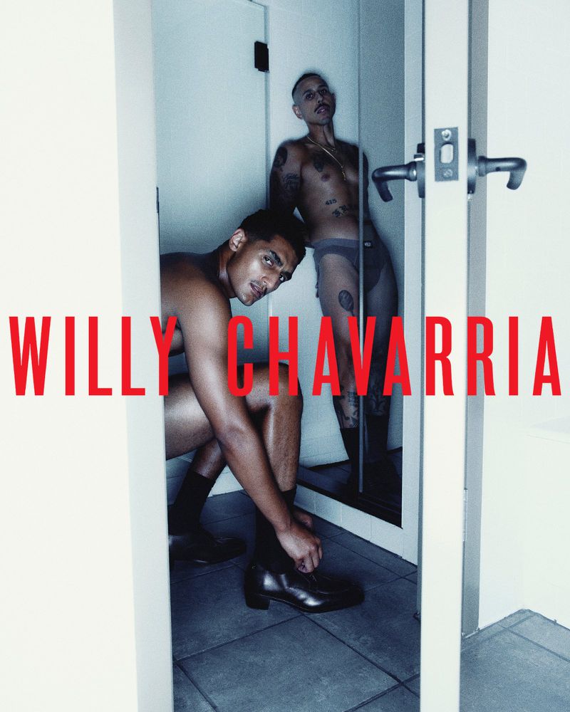 Two bare-chested men pose indoors. One man sits and ties his dress shoe, the other stands in a doorway in the background.