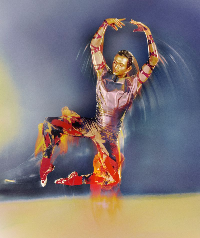 Male ballet dancer in mid-air pose with arm ribbons, striking a dynamic pose against a hazy blue backdrop. Motion blur emphasizes movement.