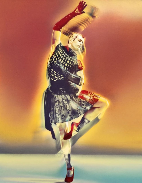A person in motion, wearing a dress, gloves, and red heels, stands on one leg against a warm-toned background, creating a blurred effect.