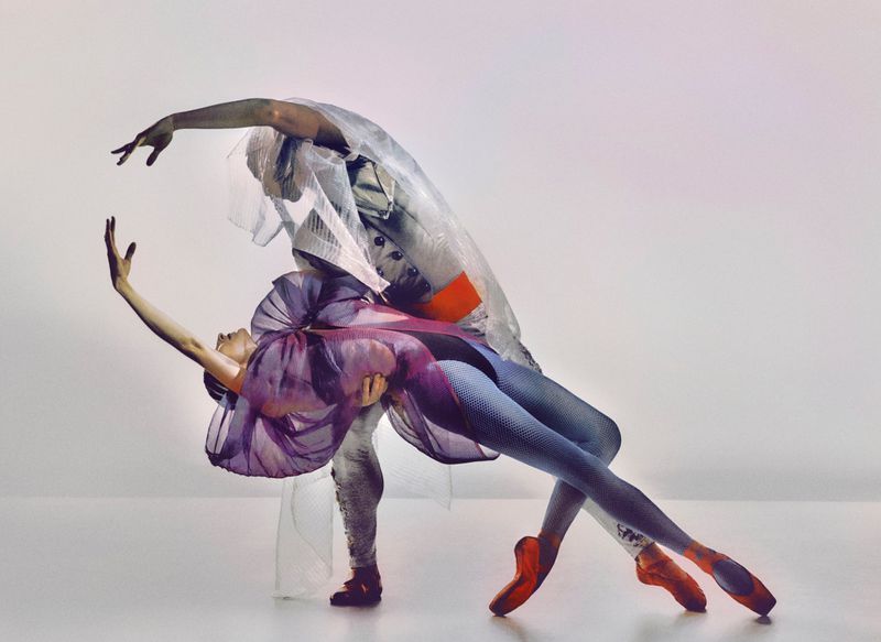 Two ballet dancers, one female and one male, are in a studio dancing in costume, the female in a purple and the male in a white costume.