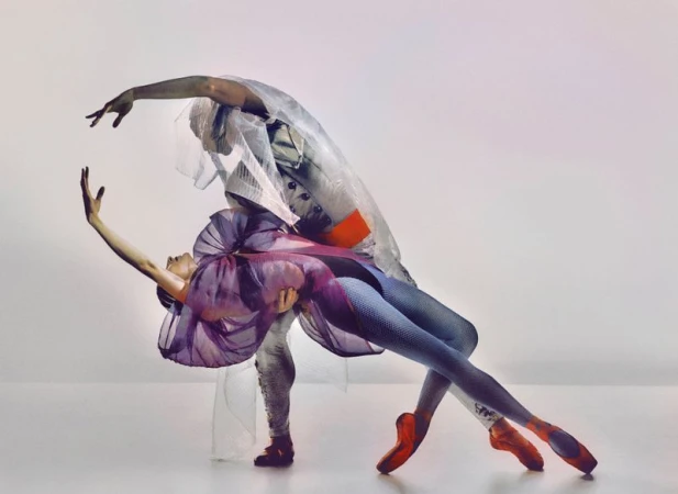 Two ballet dancers, one female and one male, are in a studio dancing in costume, the female in a purple and the male in a white costume.