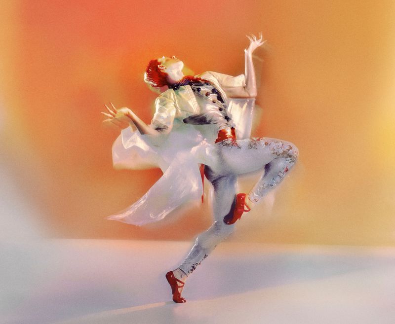 Artistic motion shot of a redhead woman in ballet pose with red shoes, floral leggings, and a white top against orange background.