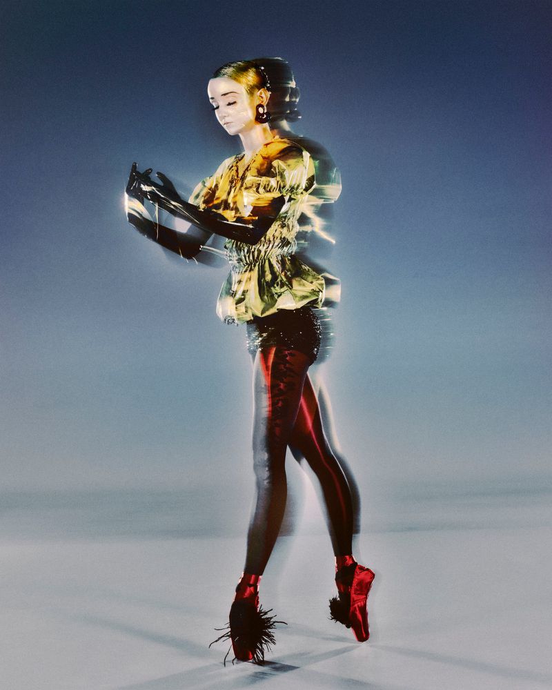 Woman in a yellow patterned top and black gloves strikes a ballet pose with red pointe shoes. Blurry studio background with soft lighting.