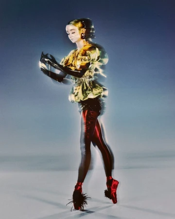 Woman in a yellow patterned top and black gloves strikes a ballet pose with red pointe shoes. Blurry studio background with soft lighting.