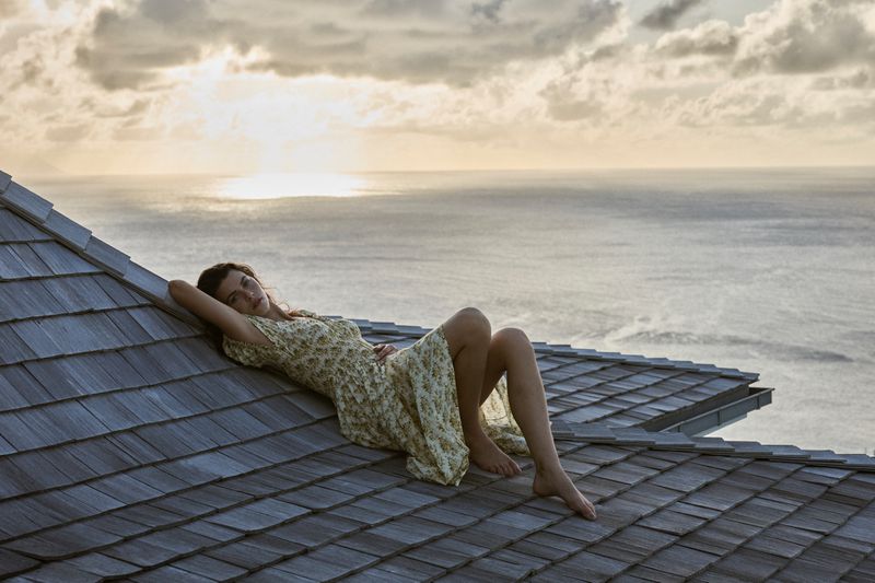 Woman in a floral dress poses on a gray shingled roof with the ocean and sky at sunset in the background, creating a serene outdoor fashion scene.
