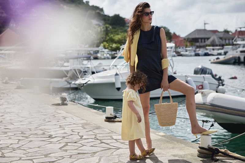 Woman in dark dress holding a straw bag standing with a child by a harbor filled with boats