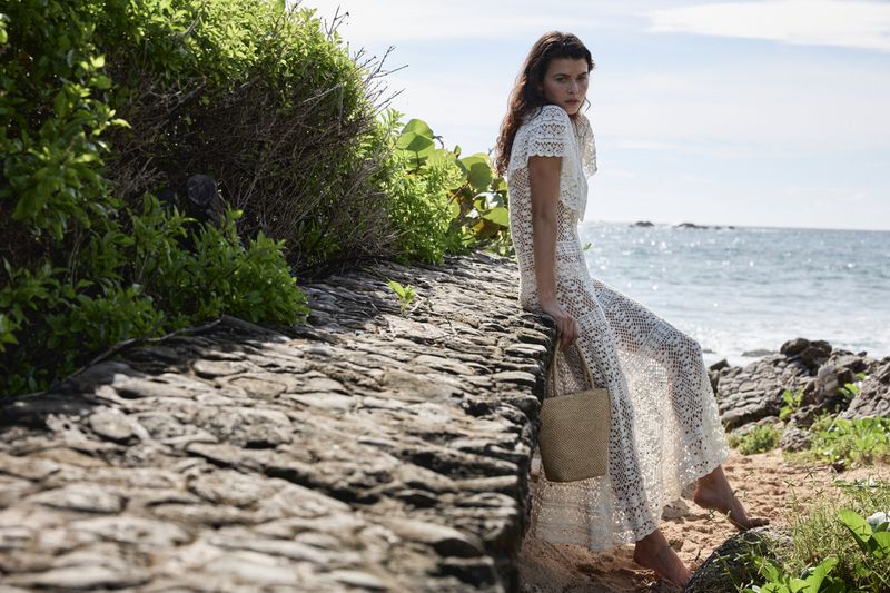 Adult female in a long white lace dress and straw bag sits on a rock wall near a tropical beach with green foliage and blue sky.