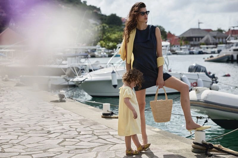 Woman in dark dress holding a straw bag standing with a child by a harbor filled with boats