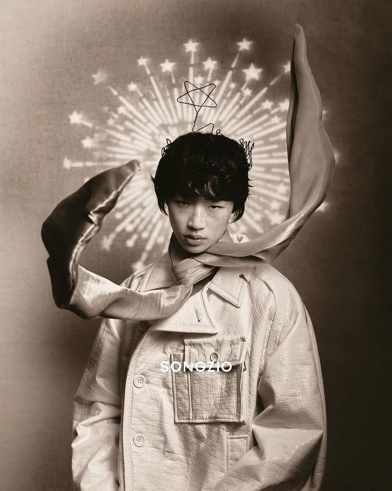 Portrait of Asian adult male wearing star crown, textured coat, and scarf against a bokeh star background in a neutral-toned photo studio.