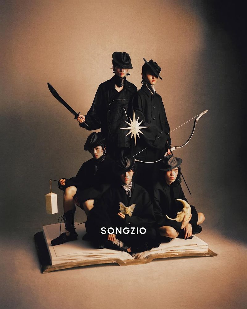 Group of five people in black attire posed on an open book, holding objects like a sword, lantern, and moon sculpture under studio lighting.