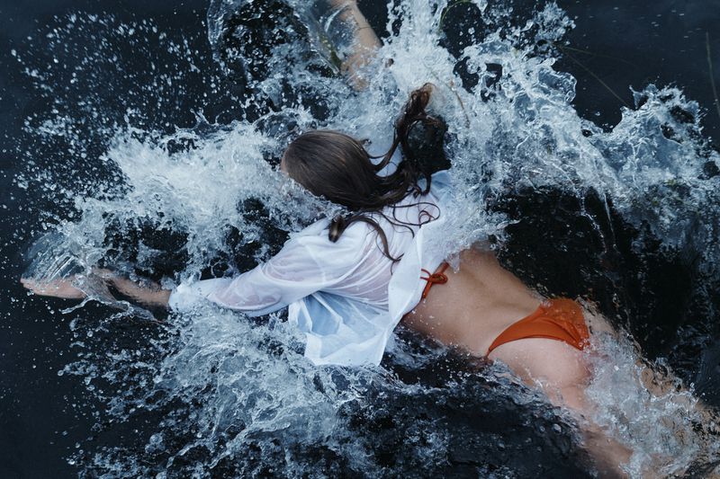 Woman in an orange swimsuit and white shirt is partially submerged in dark water, creating a splash with her arm movements.