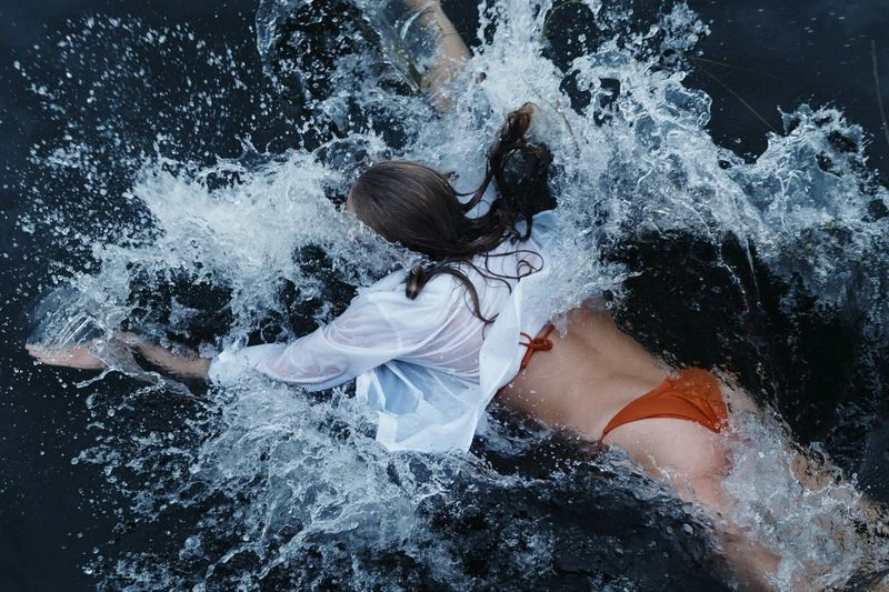 Woman in an orange swimsuit and white shirt is partially submerged in dark water, creating a splash with her arm movements.