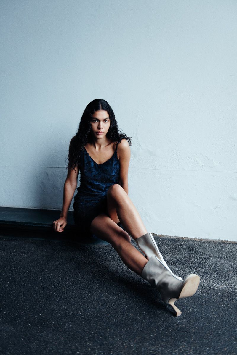 Young woman poses in a blue velvet dress and tall grey heeled boots against a grey wall on a dark grey floor in an indoor environment.