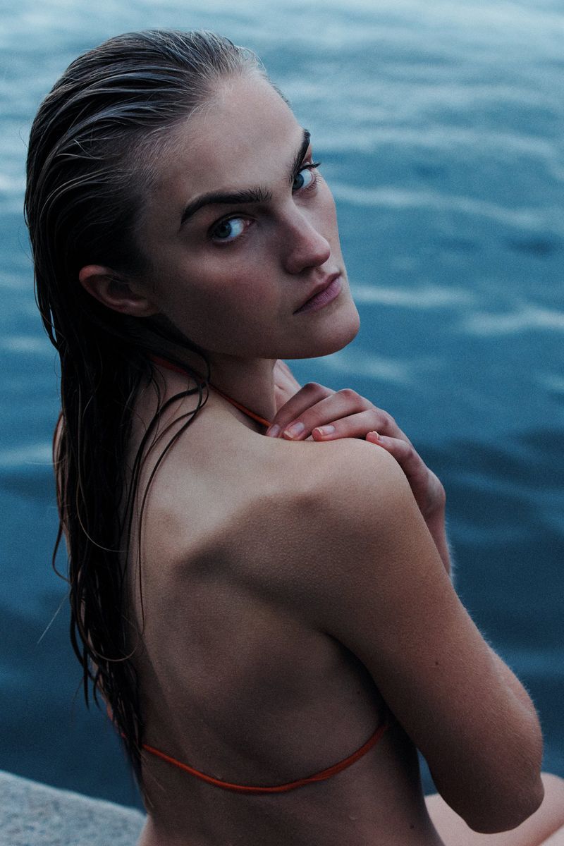 A woman with wet hair looks over her shoulder at the camera while posing outdoors against a backdrop of rippling blue water.