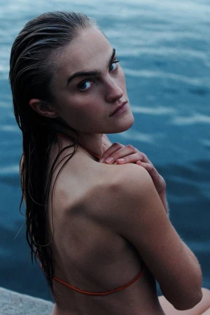 A woman with wet hair looks over her shoulder at the camera while posing outdoors against a backdrop of rippling blue water.