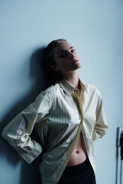 Blonde woman in unbuttoned striped shirt leaning against light blue wall. Studio portrait with low-angle, natural light, and minimalist setting.