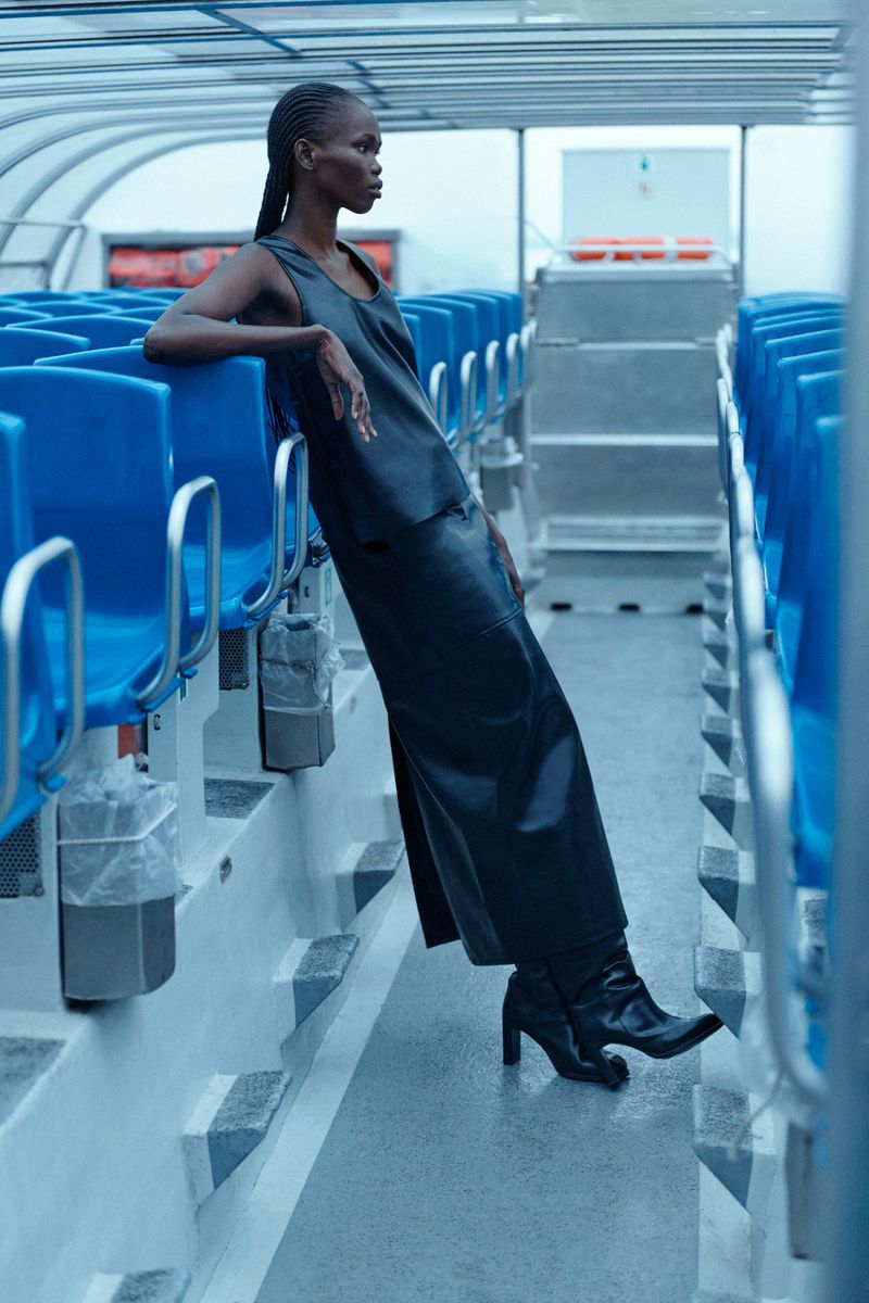 Elegant woman in a black dress and boots poses on a boat, leaning against blue seats in a fashion shot, captured in medium framing.