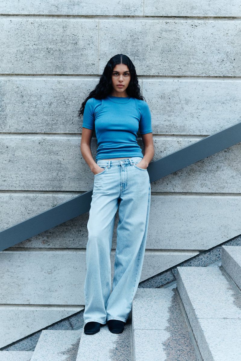 A woman poses in a turquoise t-shirt and light blue jeans against a textured concrete wall and stairs. Fashion editorial lifestyle shot.