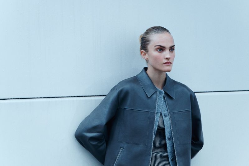 A woman poses outdoors wearing a jacket and sweater against a white wall, medium shot, cool light, fashion editorial.