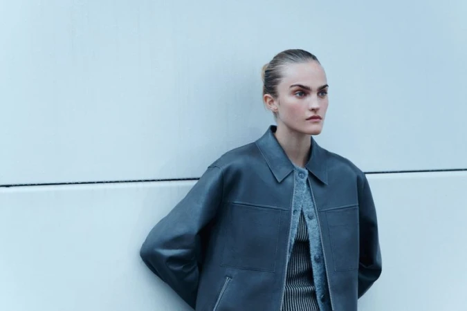 A woman poses outdoors wearing a jacket and sweater against a white wall, medium shot, cool light, fashion editorial.