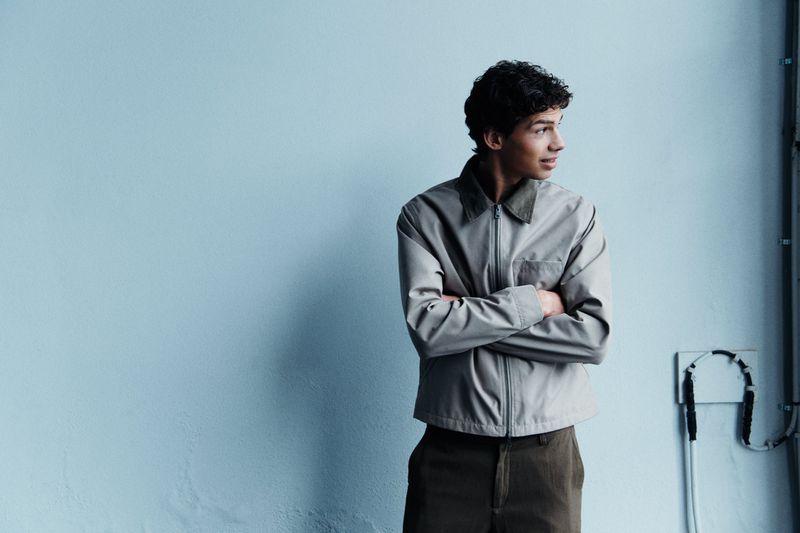 A young adult man with curly hair, wearing a light grey jacket and olive pants, stands against a pale blue wall with crossed arms.
