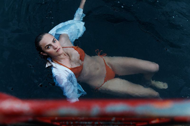 Woman in orange bikini and white shirt swims in dark water with a red railing visible at the bottom. Outdoor water fashion shoot.