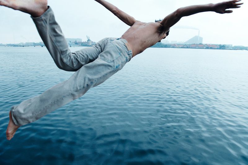 Shirtless man in jeans jumps into blue water on a cloudy day with a cityscape in the distance, captured in a wide-angle shot.