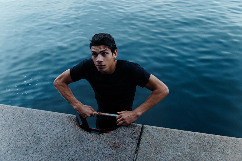 Young man in a black t-shirt emerging from blue water, holding onto a concrete ledge. Looking up and to the left, medium shot.