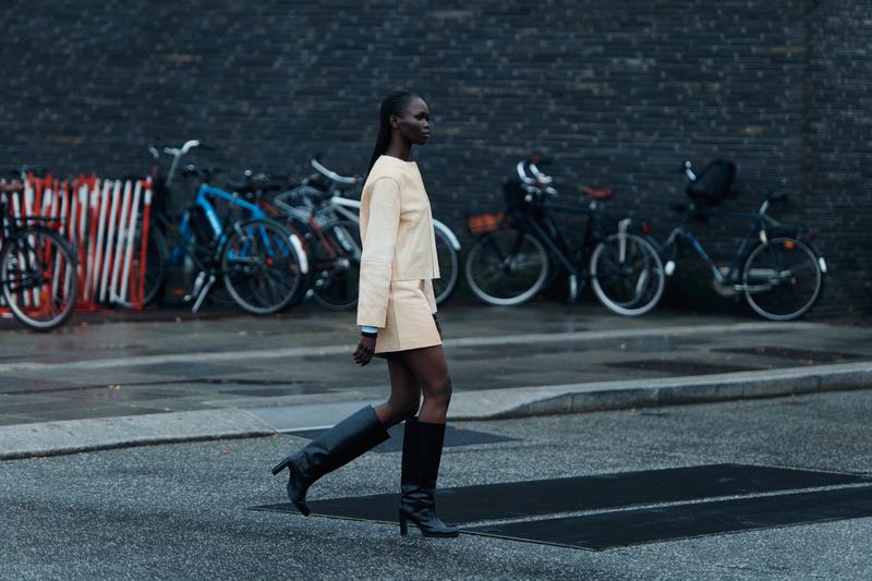 A fashion shot shows a Black woman walking on a city street in a light yellow suit and black boots, with bicycles in the background.