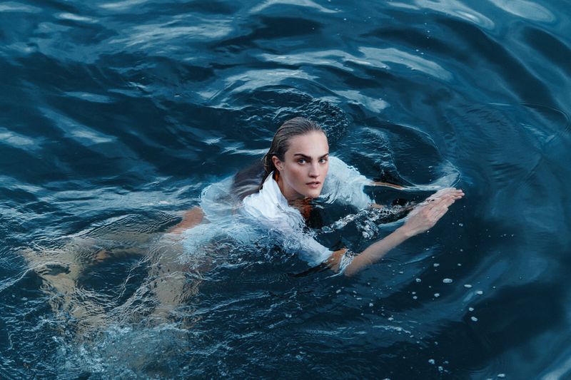 Woman swimming in deep blue water wearing a white shirt. The ocean surface has ripple effects, with dark blue tones and highlights.