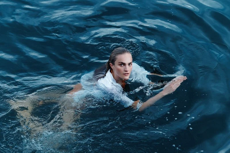 Woman swimming in deep blue water wearing a white shirt. The ocean surface has ripple effects, with dark blue tones and highlights.