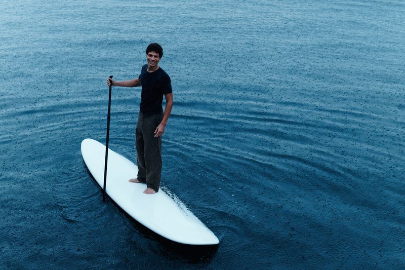 A smiling man paddleboarding on the ocean with a paddle in hand on a cloudy day with dark blue water and visible rain.