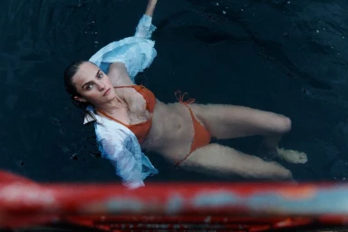 Model floating in water wearing an orange bikini and white shirt for Samsoe Samsoe Spring Summer 2025 campaign