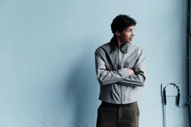 Young man with curly dark hair wearing a grey zip jacket and dark trousers standing against a pale blue wall
