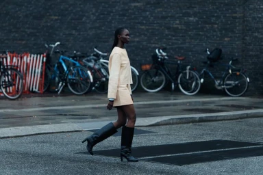 Model wearing a cream-colored top and matching skirt with black knee-high boots walking down a city street with parked bicycles.