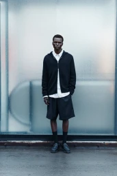 Full-length portrait of a male model wearing a black zip-up hoodie, white shirt, and black shorts standing against a frosted glass wall.
