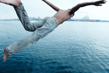 Shirtless man wearing jeans mid-air diving over open water with distant coastline under overcast sky.