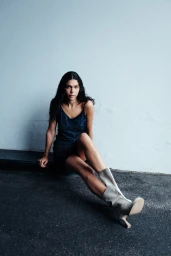 Model seated on a dark ledge wearing a dark slip dress and light boots against a wall