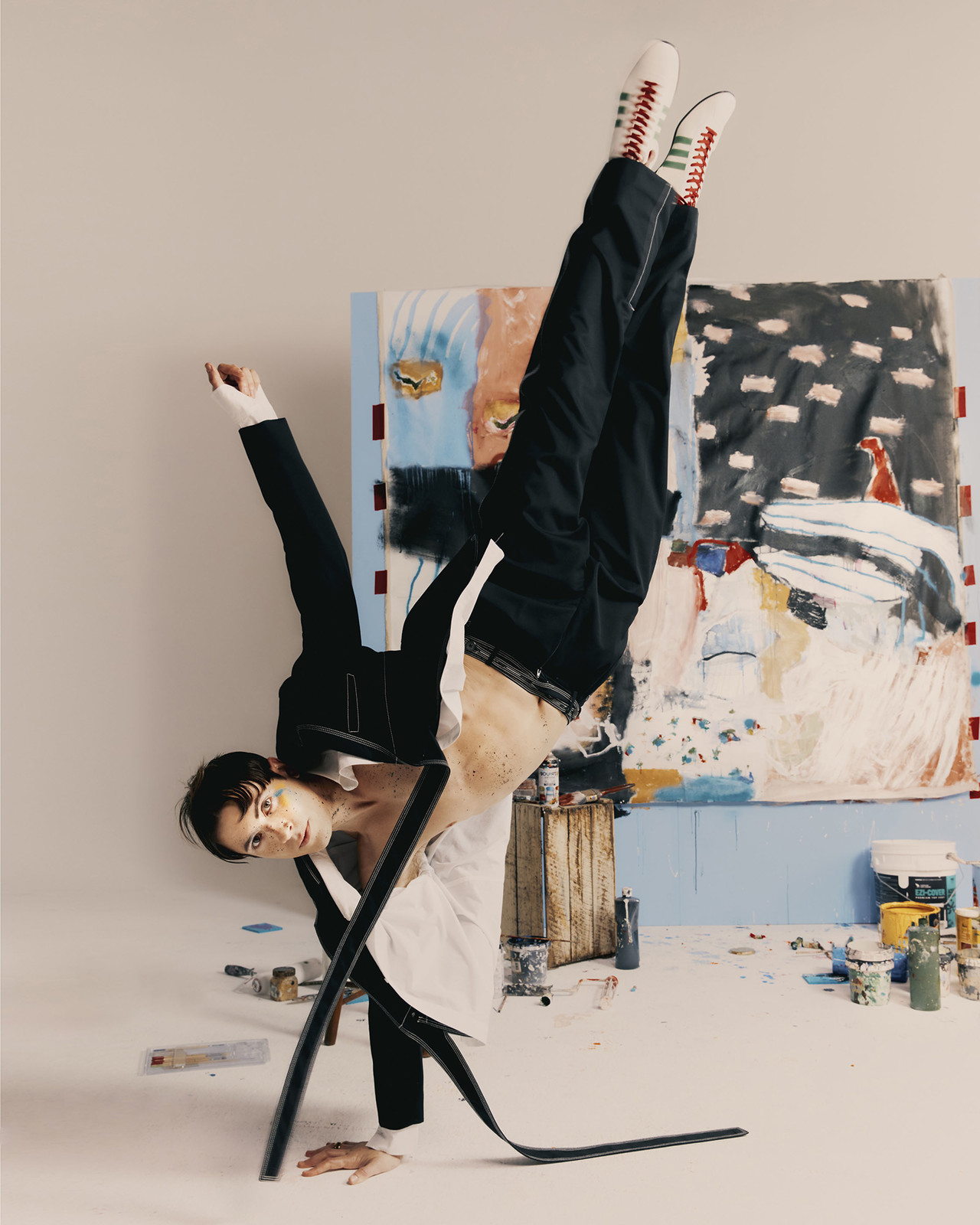 Person in avant-garde pose, wearing black pants and blazer with white shoes, in artist studio with abstract painting background.